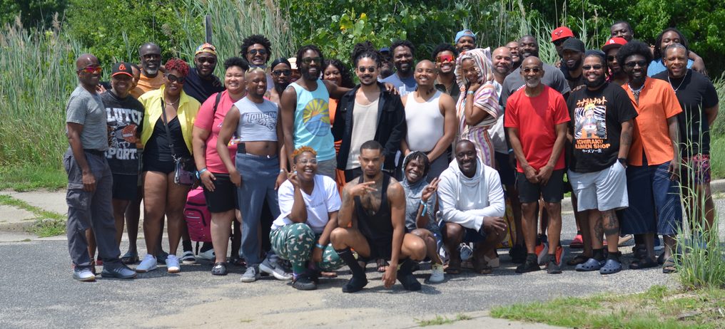 The full COURAGE Gunnison Beach group, about forty Black queer people, standing and crouching together on a road at the edge of the dunes.